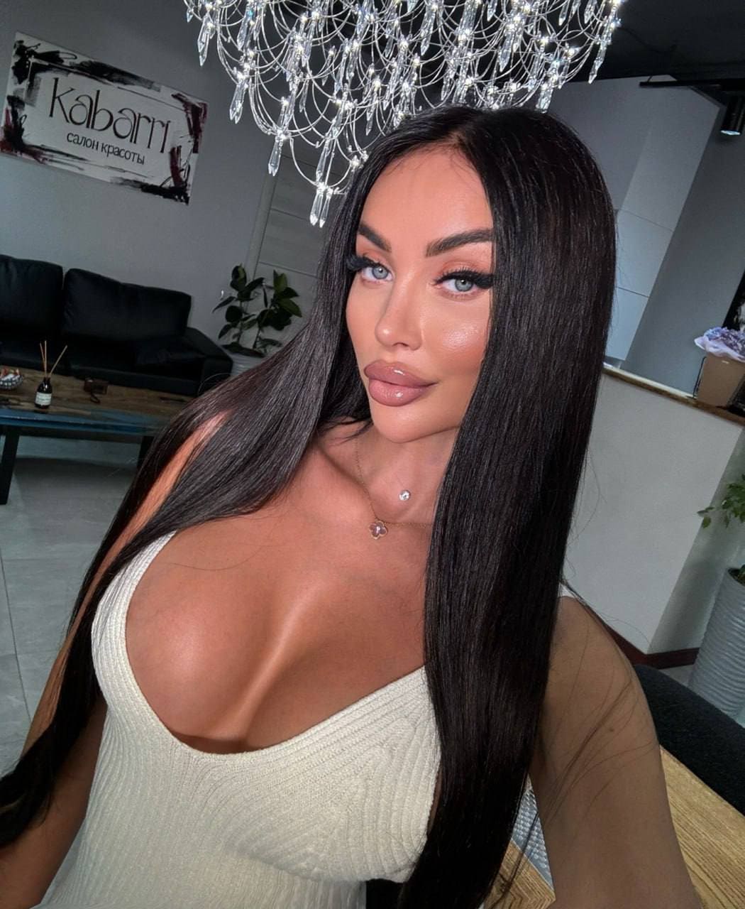 A woman with straight hair poses indoors in a white ribbed top with a deep scoop neck, beneath a crystal chandelier, with a black sofa behind her.