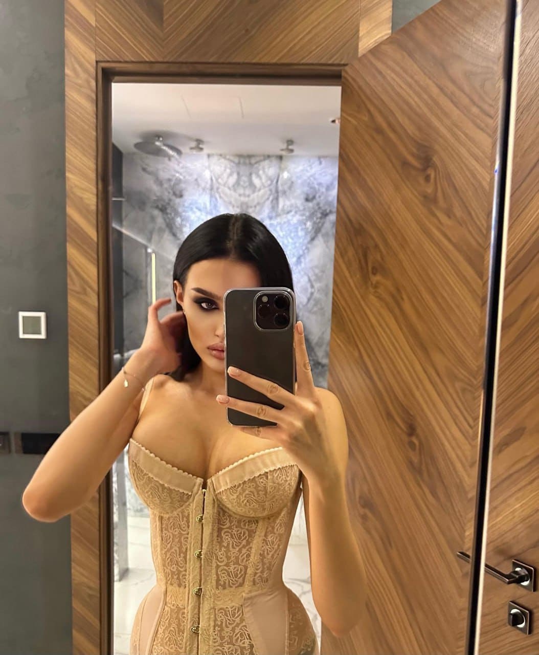 Mirror selfie in a bathroom showing a beige lace corset top with scalloped edges, one hand near the ear.